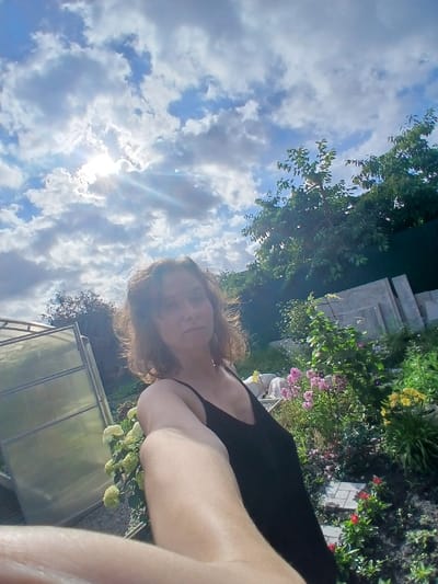 In a candid outdoor shot, a woman with shoulder-length brown hair wearing a black tank top is captured from the chest up, holding the camera in a selfie pose. She is positioned slightly to the right of the frame, her gaze directed towards the viewer. Behind her, the scene unfolds in a garden setting. To the left, a greenhouse with a translucent covering is visible. The garden bed features a variety of blooming flowers in shades of pink, yellow, and red, interspersed with lush green foliage. In the background, trees with dense green leaves and a dark green fence create a natural backdrop. The sky above is a dynamic display of blue with scattered white clouds, and the sun peeks through, casting rays of light. The overall impression is of a warm, sunny day in a cultivated outdoor space. The image may have been taken in the city of Maslova Pristan', Russia, given the context.