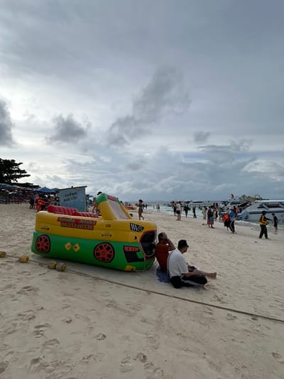 An overcast day on a sandy beach in Phru Nai, Thailand. In the foreground, an inflatable yellow and green vehicle-shaped toy with red wheels is secured by a rope and floats. Two men are sitting on the sand behind the inflatable toy. In the middle ground, a diverse group of people are walking and standing on the beach, with several speedboats moored in the shallow water. In the background, a line of beach chairs and umbrellas are visible, along with more speedboats and a dense cluster of trees. The sky is filled with heavy, grey clouds.