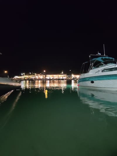 The image depicts a nighttime scene at a harbor in Limenas Markopoulou, Greece. In the foreground, a white and teal motor yacht is docked, its hull partially submerged in the dark green water. Reflections of the yacht and distant lights shimmer on the water's surface. In the middle ground, a row of illuminated buildings lines the waterfront, featuring shops and restaurants with warm interior lighting. Several other boats are moored along the quay. A yellow buoy is visible in the water near the center of the image. The background is dominated by a dark night sky. There are no people visible in the image.