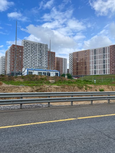 The image shows a view in Novodrozhzhino, Russia, featuring tall apartment buildings under a partly cloudy sky. A multi-lane highway with a yellow painted line on the road shoulder is visible on the right, separated from a grassy slope by a metal guardrail. The apartment buildings are primarily white and brown. One building has an antenna on top.