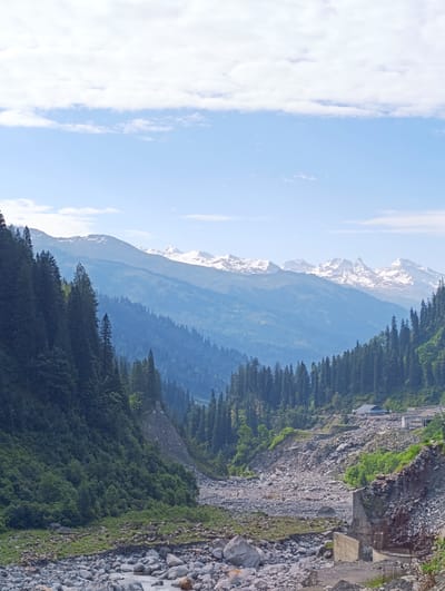 The image depicts a serene mountain landscape with snow-capped peaks in the background. The foreground is dominated by a lush green forest on the left and a rocky riverbed on the right. The sky is a clear blue with scattered white clouds, suggesting a pleasant day. The overall scene conveys a sense of natural beauty and tranquility, characteristic of mountainous regions. The presence of buildings in the distance indicates a small settlement or infrastructure within the natural environment.