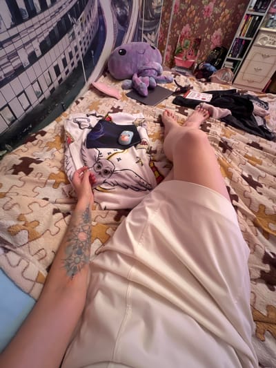 The image depicts a person lying on a bed with a puzzle-piece patterned blanket. The person's legs are extended, and they are wearing a light-colored dress or skirt. Their left arm, adorned with a tattoo, rests on the blanket. In the foreground, a white garment with a cartoon owl print is visible, along with a small toy. In the background, a purple stuffed octopus is placed on the bed. Further back, a bookshelf filled with books and a dresser are visible, along with a floral patterned wallpaper. A large mural of a modern building with many windows is on the wall behind the bed. This scene is likely in a bedroom.