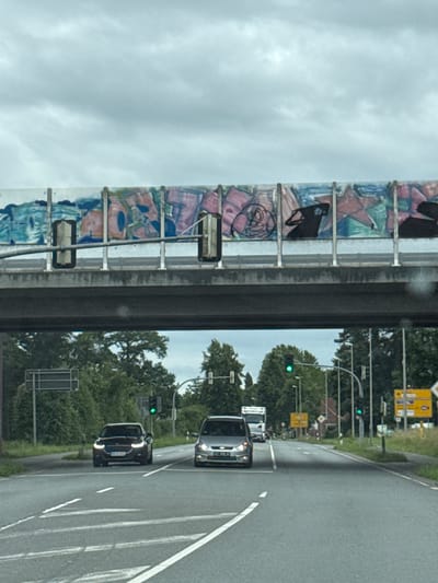 The image depicts a view from a vehicle traveling on a road in Gronau, Germany, under a cloudy sky. An overpass, adorned with graffiti featuring abstract shapes and figures, spans the roadway. Below the overpass, several vehicles are visible, including a dark-colored SUV in the foreground, a silver sedan, and a white truck further down the road. Traffic lights, some illuminated green, are positioned along the roadside, which is lined with trees and vegetation. Road signs, including directional signs, are also present. The scene appears to be captured during daytime.