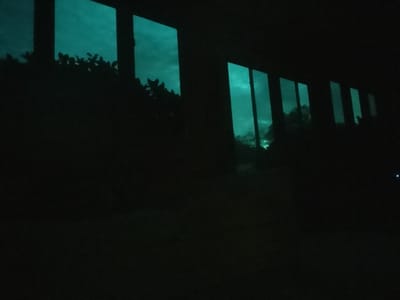 The image captures an interior view looking outward through a series of rectangular windowpanes. The scene is characterized by extremely low light, rendering the immediate interior space almost completely dark and featureless. No individuals or specific objects are discernible within the interior.
Through the multiple windows, an external environment is visible, dominated by a distinct greenish-blue illumination of the sky, suggesting twilight or an unusual atmospheric condition. Dark, indistinct masses of foliage or trees are silhouetted against this light, visible through the lower and mid-sections of the window views. Dark, vertical and horizontal bands within the frame delineate the individual windowpanes, consistent with window sashes or architectural divisions. A subtle, brighter greenish glow appears near the base of some distant window views, potentially indicating a light source or reflection in the external environment.
The location is stated to be in Roswell, United States; however, no specific landmarks or identifying features within the visual content of the image allow for a precise determination of its exact location within the city. No actions or interactions are depicted.