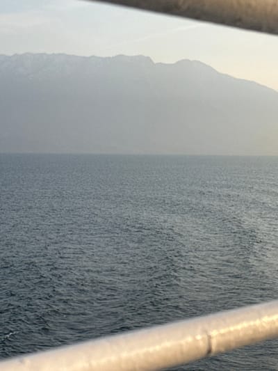 The image depicts a panoramic view of a body of water with a mountain range in the background, observed from what appears to be a vessel or elevated platform in Kato Karioutes, Greece. In the immediate foreground, a light-colored, metallic handrail extends diagonally from the lower left towards the lower right, partially obstructing the view of the water. Above this, the midground is dominated by a vast expanse of calm, dark blue-grey water exhibiting minor surface ripples. The distant background features a lengthy, undulating mountain range stretching across the horizon. These mountains appear muted and indistinct, suggesting significant atmospheric haze or distance. The sky above the mountains is a pale, uniform grey-blue, indicative of overcast conditions or diffused light. A thin, dark, horizontal bar or structure is visible at the very top edge of the frame, further segmenting the sky. No individuals, additional vessels, or specific architectural elements of a city are visible within the frame, beyond the general environmental features.