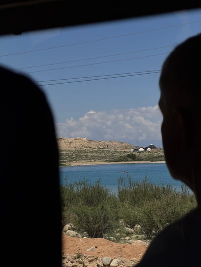 This image captures a serene view of a body of water, likely Issyk-Kul Lake, near Tong, Kyrgyzstan, as seen from inside a vehicle. The foreground is dominated by the silhouetted heads of two individuals, suggesting they are passengers. The primary subject of the image is a vast, calm blue lake, with a distant, rugged shoreline characterized by sandy hills and sparse vegetation. On the far bank, several structures, including traditional yurts and modern buildings, are visible, hinting at a settlement or resort. In the middle of the lake, a small inflatable raft with two occupants can be seen, adding a touch of human activity to the landscape. The sky is a bright blue, dotted with fluffy white clouds, and several power lines stretch across the frame, indicating the proximity of infrastructure. The overall impression is one of natural beauty and a moment of peaceful observation.