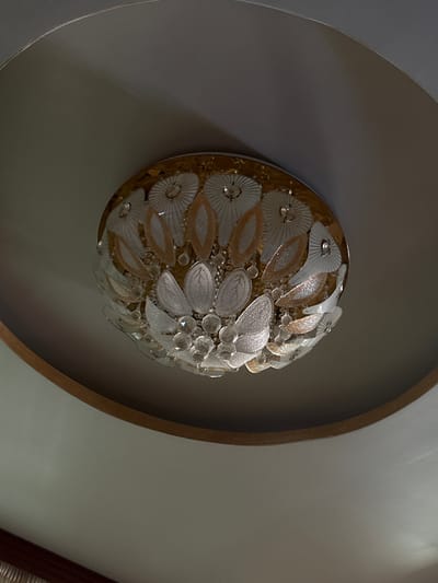 The image shows a close-up of a decorative glass ceiling light fixture. The fixture is spherical and appears to be made of a mosaic of glass elements resembling petals and leaves, with some crystal accents. The colors are predominantly white and gold. The fixture is mounted on a ceiling that has a circular, recessed border, painted in a light gray color. The lighting in the image is dim, with shadows cast on the ceiling, suggesting it might be nighttime or the fixture is not currently illuminated. There is no visible text in the image. The context provided places this scene in Slanchevo, Bulgaria.