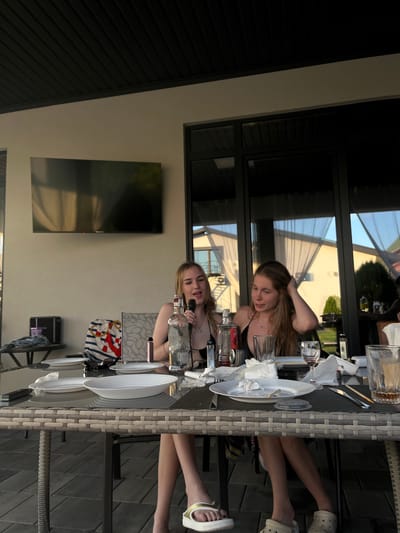Two young women are seated at a table on a patio. The woman in the foreground, with blonde hair, is wearing a black bikini top and looking at the camera with a slight smile. She is holding a bottle of what appears to be alcohol. Beside her, another young woman with blonde hair, also wearing a black top, is partially visible. They are seated at a woven outdoor table set with plates and glasses. In the background, a large screen television is mounted on a light-colored wall. The scene appears to be outdoors during the daytime, with sunlight reflecting off the glass doors and windows. The overall impression is a casual gathering on a warm day.