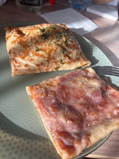 The image shows a plate with two folded pancakes or crepes, possibly in Ladozhskii Trudposelok, Russia, given the prompt. A fork rests between them. The crepe on the left has a golden-brown surface speckled with green herbs, while the crepe on the right is lighter with a pinkish-red filling or topping. The plate has a light-colored surface with a repeating square pattern. A bottle and a napkin are visible in the background, along with a wooden surface. The lighting is bright, with strong shadows.