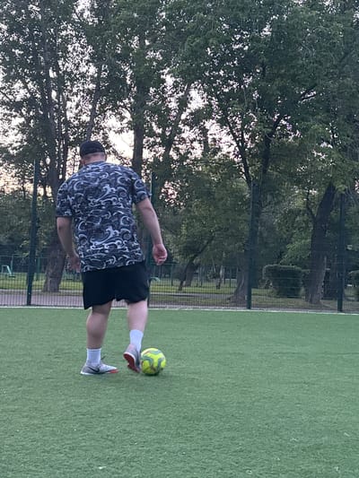 A person is playing soccer on an artificial turf field. The person is wearing black shorts, a dark t-shirt with a white swirl pattern, white socks, and athletic shoes. The person is kicking a soccer ball, which is yellow and green. The field is bordered by a fence and trees. The time of day appears to be late afternoon or early evening, as indicated by the soft lighting and shadows. The weather seems to be mild, with no visible precipitation or strong winds. The location is Petropavl, Kazakhstan. There are no visible text elements in the image.