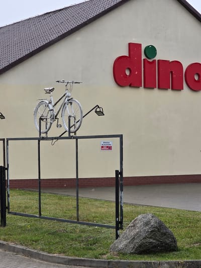 A white bicycle is mounted on a metal structure in front of a Dino supermarket. The supermarket's logo, a red "dino" with a green circle, is visible on the building's facade. A "DRZWI WJAZDOWE ZAKAZ PARKOWANIA" (Entrance Road No Parking) sign is attached to a fence. The scene is located in Trąbki Wielkie, Poland.