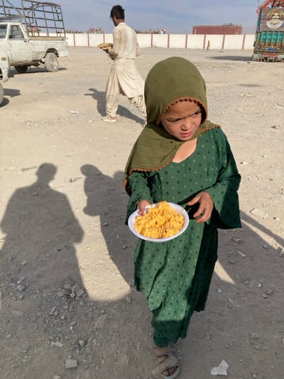 An outdoor scene in Chaman City Tehsil, Pakistan, depicts a young girl and an adult male in an unpaved, dusty environment under daylight.
In the foreground, a young girl, approximately 4-7 years old, is centered, holding a white disposable plate containing orange-yellow rice. She wears a dark green full-length dress with a subtle pattern and an olive-green head covering trimmed with an orange-brown crocheted edge around her face. Her expression is neutral, and her gaze is directed slightly downwards and to her left. She wears light-colored sandals.
In the mid-ground, behind the girl and to her right, an adult male walks away from the camera. He wears light-colored traditional clothing (shalwar kameez) and carries a flat, round item, possibly bread or another food item, in his left hand.
The ground is unpaved, consisting of dirt and gravel, with visible shadows of at least two individuals, including the photographer, extending into the foreground.
The background features a low, light-colored concrete wall running horizontally. Behind the wall, a white pickup truck with a metal frame is parked on the left, and a colorfully decorated truck is visible on the far right. A dark shipping container is also present. Several distant, blurry figures are visible walking near the wall and container. The sky above is light blue with sparse cloud cover.