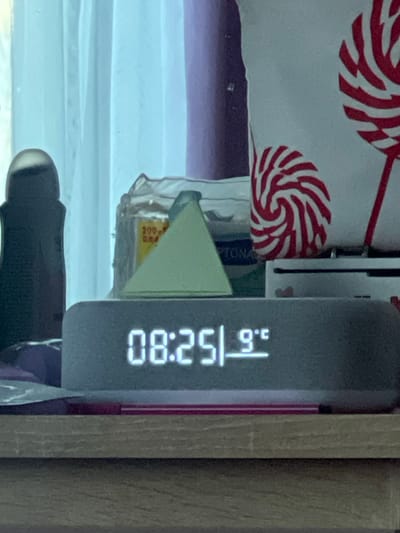 A digital clock displays 08:25:80, with a temperature reading of 23.6°C. The clock's grey case sits near a wooden surface. Behind it is a white cloth with red and white swirl patterns. A triangular green object and a package labeled "200 GRAM" are partially visible near the clock. The background includes glimpses of a window with horizontal blinds and assorted items suggesting a domestic setting.