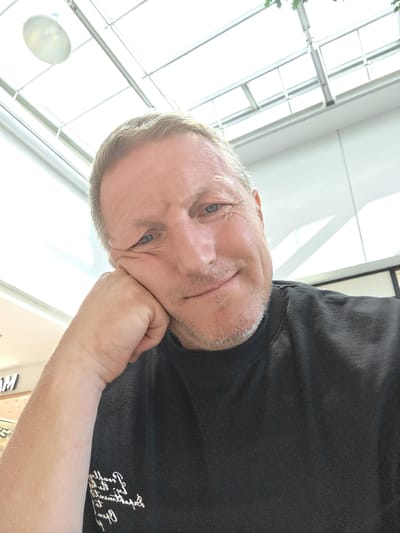 A man in his late 40s or early 50s with short, light brown hair and blue eyes rests his head on his hand, looking directly at the camera. He has a faint stubble and is wearing a black t-shirt. The background is out of focus but appears to be an indoor setting with a bright, reflective ceiling that has a grid-like structure, possibly a mall or a modern building with a skylight. The image is taken from a low angle, looking up towards the man. There are no discernible landmarks that would place this scene in Nola, Italy.