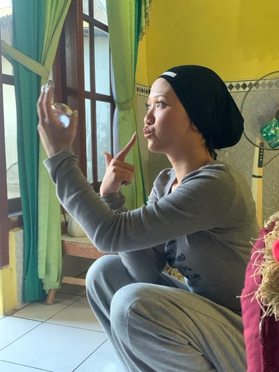 A young woman wearing a black hijab and a grey tracksuit is crouched by a window with green curtains. She is looking to her right, with her mouth pursed, and holding a small, reflective object in her left hand, while pointing with her right index finger. The room has yellow walls and tiled floors. In the background, a fan is visible. The scene appears to be indoors in a residential setting in Jajag, Indonesia.