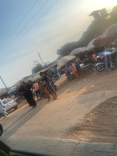An outdoor scene depicts a bustling street market alongside a road. Several individuals are gathered, likely shopping or selling goods under umbrellas and makeshift stalls. A motorcycle is visible among the market items. The road surface appears worn, and power lines stretch across the clear, bright sky. The scene suggests a vibrant, active community in a tropical region.