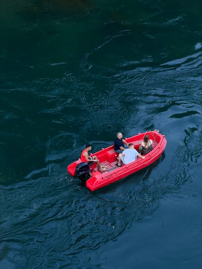 Four individuals are aboard a bright red inflatable boat with an outboard motor. The boat is positioned on dark, choppy water. In the stern, a person wearing a red tank top and black shorts operates the boat. Ahead of them, three passengers are seated. The individual in the front passenger seat wears a light blue t-shirt and grey shorts. The person next to them is wearing a dark blue t-shirt and jeans. The fourth person, a woman, is seated further back and wears a patterned dress. The boat appears to be moving, as indicated by the ripples in the water. This scene is located on the Neretva River in Mostar, Bosnia and Herzegovina, likely near the iconic Old Bridge, given the common tourist activities in that area. The water's deep, dark color suggests depth and possibly strong currents. The overall impression is one of leisure activity on a river.