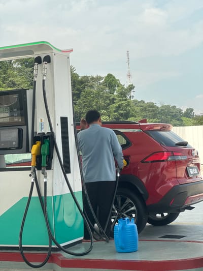 A person is fueling a red car at a gas station. The person is wearing a light blue long-sleeved shirt and is holding a yellow fuel nozzle. The car has a license plate that reads "BRU 5500". A blue plastic jug is visible next to the car's rear wheel. The gas pump has green and yellow nozzles. The scene appears to be during the day, with overcast skies. In the background, there are trees and a tall radio tower. The gas station has a white and green color scheme. Visible text on the gas pump includes "PETRONAS PRIMAX 95 with Pro-Drive". The location context indicates Subang Jaya City Council, Malaysia.