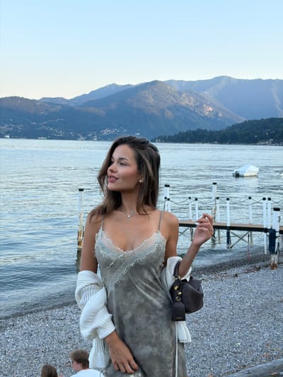 A young woman stands on a pebble beach near a lake in Nobiallo, Italy. She is wearing a light grey slip dress with lace detailing, a white cardigan draped over her shoulders, and a dark brown handbag. The lake is calm, with a few boats visible in the distance. Mountains form the backdrop, with a clear blue sky overhead. Two other individuals are partially visible in the lower left corner of the frame, seated on the beach.