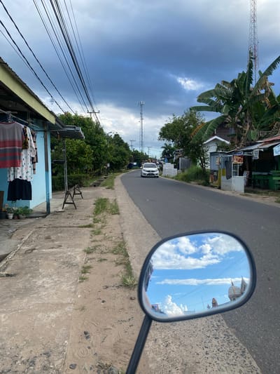 The image captures a street scene in Sungai Sipai, Indonesia, viewed from the perspective of someone riding a motorcycle. A light-colored car is driving on the asphalt road, which curves to the right. On the left side of the road, a light blue building with a yellow overhang is visible. Clothes are hanging on a line in front of the building. A sidewalk made of concrete and dirt runs alongside the building, with some patches of grass growing through it. Further down the road, trees line the sides, and various structures, including what appear to be small shops and houses, are present. A tall telecommunications tower is visible in the distance. The sky is cloudy with patches of blue sky. A motorcycle's side mirror in the foreground reflects the sky and some building structures.