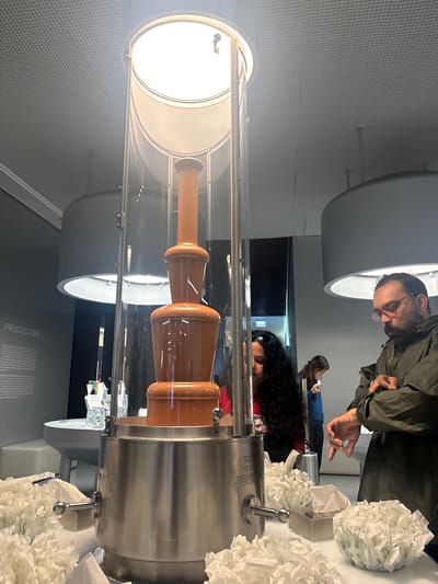 A vertical, medium-shot indoors depicts a chocolate fountain encased in a transparent tube, with two people in close proximity. The fountain, filled with a cascade of flowing brown chocolate, has a built-in light source on the upper part of the tube. To the right of the fountain, there is a metallic spigot, accompanied by a generous pile of white paper napkins. The environment indicates that the location is inside a museum or a shop, possibly in Kilchberg (ZH), Switzerland. 

One individual, seen from the waist up, has dark hair, is wearing sunglasses, and is located just behind the fountain, as if viewing the process. Another person, also seen from the shoulders up, wearing glasses and an olive green jacket, appears to be pointing or gesturing towards the fountain. Behind this individual, another person is seen in the distance, further back in the environment. The lighting is bright, highlighting the chocolate fountain as the central focus of the shot.