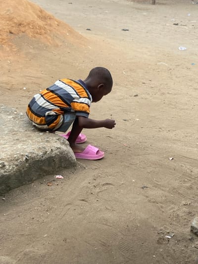 The image displays a young child, appearing male, with dark skin and short dark hair, squatting outdoors on a concrete step in Ojo, Nigeria. The child wears a horizontally striped short-sleeved shirt in white, light grey, dark grey, and orange, paired with dark blue shorts and bright pink slip-on sandals. The child is leaned forward, looking intently at the light brown dirt ground, with their right hand lowered as if examining or interacting with a small item. The immediate environment consists of sandy-dirt ground with visible tire tracks, shoe prints, and scattered small pieces of litter. A distinct white wrapper lies on the ground near the concrete step. In the upper left background, a large mound of light brown earth or sand is visible. The scene is unpaved and appears to be an open, unkempt area.