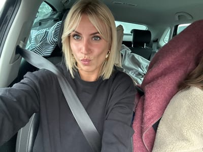 An image taken inside a vehicle in the city of Brignoles, France, shows two individuals. A blonde female, with shoulder-length hair and bangs, occupies the driver's seat. She is wearing a black long-sleeved sweatshirt, gold-colored hoop earrings, and a grey seatbelt diagonally across her chest. Her lips are pursed, and she looks directly at the camera. To her right, a second individual is partially visible in the passenger seat, wearing a light-colored, possibly cream-toned, fleece jacket, with a maroon or burgundy garment draped over them. The backseat is visible behind the driver, containing luggage including a black bag with a striped garment and a grey hooded sweatshirt. The car interior features dark upholstery with white stitching on the seats.