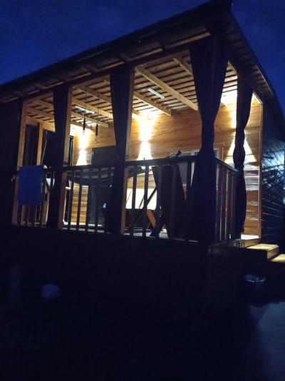 The media file depicts a wooden gazebo or porch attached to a wooden building, illuminated by warm, interior lighting. The scene is set at night, as indicated by the dark blue sky and the artificial illumination of the structure. The gazebo features a slatted wooden roof and is supported by wooden pillars. Dark curtains hang down on either side of the open front of the gazebo, adding a sense of enclosure. A wooden railing encloses the porch area.
Visible objects within the gazebo include what appears to be furniture, possibly chairs and a table, though they are partially obscured by shadows and the curtains. A blue towel is draped over the railing on the left side of the porch. There are also light fixtures visible on the ceiling of the gazebo. The ground in front of the gazebo appears to be dark and possibly covered in grass or gravel. Steps leading up to the porch are visible on the right side of the frame.
The overall mood is tranquil and inviting, suggesting a relaxing evening setting. There are no people visible in the image. The location cues, such as the style of the building and the natural surroundings, are consistent with a residential or recreational area in Degtyarsk, Russia. No visible text is present in the media file. The lighting creates strong contrasts, with bright yellow light emanating from within the gazebo against the dark night sky and the silhouetted foreground.