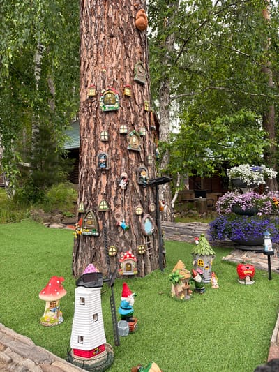 This is a forensic, matter-of-fact description of a garden scene in Sukhaya, Russia. The image depicts a large tree trunk adorned with numerous small, decorative houses and windows, creating a whimsical fairy tale aesthetic. These miniature structures are affixed to the bark at various heights and angles.
At the base of the tree, the ground is covered in artificial turf. Several garden ornaments are scattered across the lawn. To the left, there is a red and white mushroom-shaped house with a smaller mushroom on top. Next to it stands a white lighthouse-shaped structure with red accents and black window openings. A garden gnome in a blue outfit and red hat is positioned to the right of the lighthouse, holding a silver bucket.
Further to the right, a cluster of fairy houses is visible. One is a tall, green, fairy-tale style house with a yellow window. Another is a smaller, tan, two-story house with a wooden balcony. Two garden gnomes are standing near these houses, one taller gnome in green and a smaller one in pink. To the far right, a red apple-shaped house with "Fruit Shop" written on it is present.
In the background, behind the tree and garden ornaments, there is a wooden house with a green roof. Beyond that, lush green trees and foliage form a dense backdrop. To the right of the tree, there are tiered planters overflowing with colorful flowers: white and blue flowers in the top planter, pink flowers in the middle, and vibrant purple flowers in the bottom planter. A small lantern-style light fixture is visible to the far right. The overall environment is a well-maintained outdoor garden space. There are no people visible in the scene.