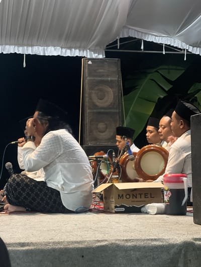 In Wedarijaksa, Indonesia, a group of men dressed in white attire and black skullcaps are seated on a stage under a white tent. The man in the foreground, with long dark hair and a black skullcap, is holding a microphone and appears to be singing or speaking. He is wearing a white, intricately patterned shirt and black patterned pants. To his right, four other men are also seated, three of whom are holding tambourines. Behind them, a large speaker is visible, along with what appears to be banana leaves from the environment. In front of the group, a cardboard box with "MONTEL AIR MINERAL" printed on it and a large jug of dark liquid with a pink lid are placed on the ground. The scene suggests a performance or a gathering with musical accompaniment.