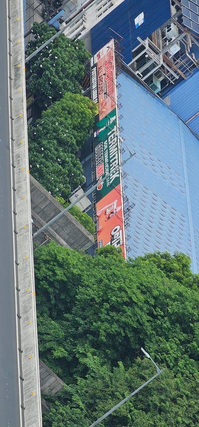 The image, captured in Makati, Philippines, shows a street-level view of a billboard advertising "Solignum" and "TERMITRAK SOIL TERMITEICIDE." The billboard is prominently displayed alongside a building with a blue, corrugated metal roof. Lush green trees, some with white flowers, surround the base of the billboard and the building. A concrete overpass or elevated road structure is visible in the foreground, with yellow markers along its edge. In the background, parts of other buildings and industrial equipment are visible. The scene suggests an urban environment with a blend of commercial structures, natural elements, and infrastructure.