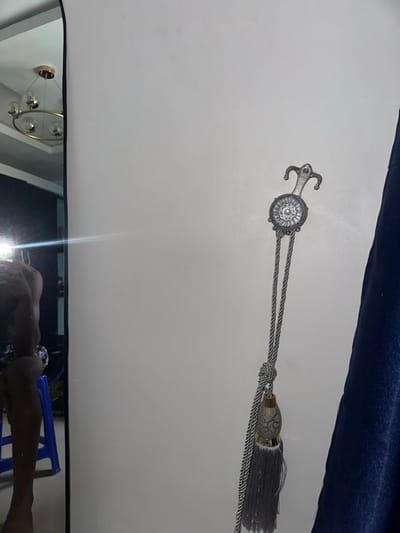 The image depicts an indoor scene in the city of Obinze, Nigeria, primarily featuring a plain, light-colored wall. On the left, a tall, vertical mirror reflects portions of a room. Within the mirror's reflection, a person, appearing to be male, is visible from the mid-torso to mid-calf, positioned with their back partially towards the mirror. A bright light source is reflected in the mirror, obscuring the upper part of the person's reflection. A blue plastic stool is partially visible behind the person in the reflection. Above, the reflection of a gold-framed, multi-globe chandelier is seen against a white ceiling. On the right side of the main wall, a decorative curtain tie-back is mounted. This tie-back features an ornate, silver-toned metallic holder with a central circular motif containing a pearl-like element. A double braided grey cord hangs from the holder, knotted in the middle, and culminates in a grey tassel with a cracked-texture oval bead. A dark blue, textured curtain is partially visible along the far right edge of the frame.