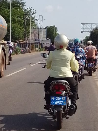The image captures a scene on a road in what is presumed to be Dungu, Ghana, given the presence of a motorcycle license plate with the country code "GH." Several motorcycles are traveling in the same direction, with the closest one to the camera displaying a license plate that reads "M-25-NR 6937." The rider of this motorcycle is wearing a pale yellow shirt and a helmet. Other motorcycles are visible ahead, each carrying a rider. The road appears to be paved and marked with lane dividers. A tanker truck is partially visible on the left side of the road. In the background, there are buildings and vegetation, as well as an advertising billboard structure. The sky is visible and appears to be clear.