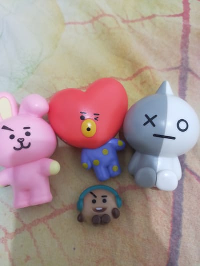 Four BT21 character figurines are arranged on a patterned cloth surface. From left to right, the figurines are: a pink rabbit with yellow accents and black markings; a red heart-shaped character with blue and yellow polka dots, featuring a yellow nose and a wink; a grey and white character with black markings resembling an X and a line for an eye; and a small, brown, round character with blue headphones and brown accents. The cloth has a mottled pattern of yellow, white, and pinkish-red lines. The image was taken indoors in the city of Pematang Siantar, Indonesia.