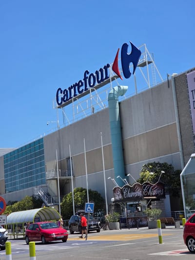 The image captures a street-level view of the Carrefour supermarket in Collado Villalba, Spain. The large blue "Carrefour" logo with a red accent is prominently displayed on the building's facade. In front of the store, a red hatchback car is stopped at a crosswalk. To the right of the crosswalk, a person in a red shirt stands next to a black SUV. A kiosk advertising "La Churreria Churros" with "Refrescos Caseros" (Homemade Refreshments) is visible to the right of the store's entrance. The sky is clear and blue, and the scene is illuminated by bright sunlight. A disabled parking sign is also visible.