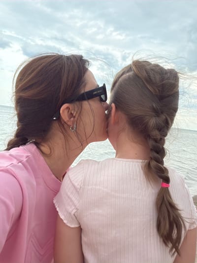 The image depicts two individuals, a woman and a child, in close proximity outdoors, likely at Plieņciems beach in Latvia. The woman is wearing sunglasses with the brand name "JIMMY CHOO" visible on the side. She is also wearing a pink sweatshirt and earrings. The child has braided brown hair with a pink hair tie. Both individuals appear to be touching faces. In the background, there is a beach and a cloudy sky.