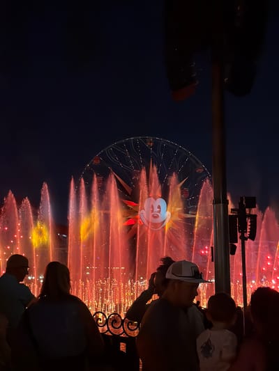 The image captures a nighttime scene at Disney California Adventure Park in Anaheim, California. The Ferris wheel, commonly known as Pixar Pal-A-Round, serves as a focal point, illuminated with a projection of Mickey Mouse. A water show occurs in front of the Ferris wheel, with streams of water lit in various colors, including red, yellow, and orange. The foreground shows people watching the show, including an adult with a child, and others standing nearby. A structure, possibly a railing, is visible between the viewer and the Ferris wheel, partly obstructing the view. The overall ambiance of the photograph is that of an evening entertainment event.