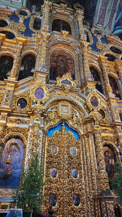 The image shows a portion of a highly ornate, gilded iconostasis. It features numerous religious paintings framed by elaborate gold carvings. The iconostasis is part of the Church of the Intercession (Pokrov Church) in Smolensk, Russia. There are no people visible in the photo, but the icons depict religious figures. The environment is inside the church, likely near the altar.