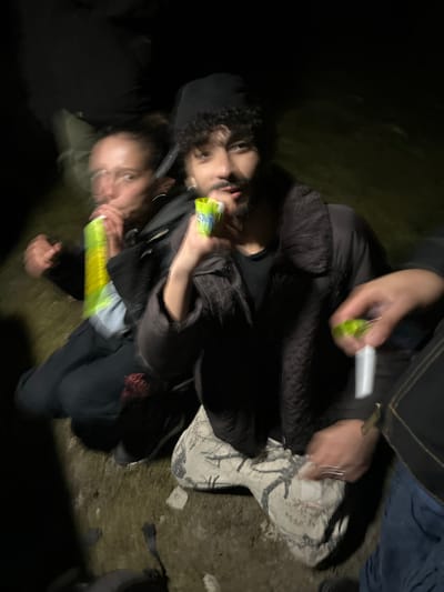 In a dark outdoor environment, three people are seen. One individual in a black hat and jacket holds a yellow object, while a second person beside them appears to be holding a similar item. A third individual, wearing a dark jacket and jeans, holds an object in their hand. The surroundings consist of ground cover and possibly a grassy area. The location is presumed to be Auvers-Saint-Georges, France.