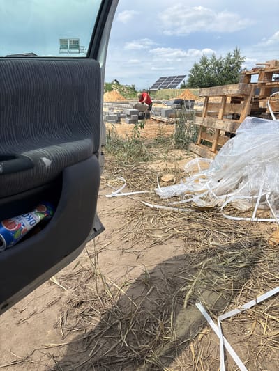 The image depicts an outdoor scene at a construction site, possibly in Opatów, Poland, under a partly cloudy sky. In the foreground, a car door is open, revealing its interior with a gray fabric-lined door panel and a beverage can in a storage compartment. The ground is covered with dry grass, straw, and dirt.
Further into the scene, construction is underway. A person wearing a red shirt is seen working with concrete blocks, possibly laying the foundation for a building. A wheelbarrow is nearby, and piles of sand and more concrete blocks are visible. In the background, solar panels are installed on a roof, and wooden pallets are stacked, suggesting construction material storage. The overall environment appears to be a developing area, with elements of both construction and rural landscape.