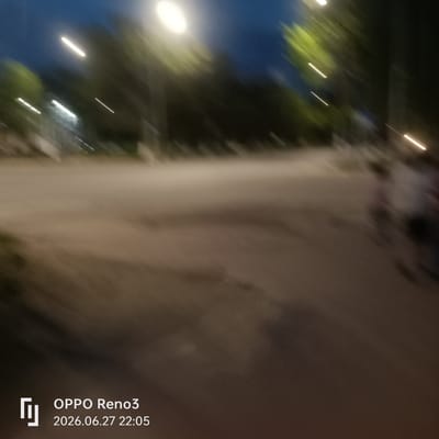 The image captures a blurred outdoor scene at dusk or night in Atyrau, Kazakhstan. The primary subjects are indistinct figures and trees, with light streaks suggesting movement or artificial lighting. The ground appears to be a dirt or gravel path, possibly a park or unpaved road, with some structures faintly visible in the background on the left, possibly buildings or streetlights.
The scene is characterized by a dark blue sky with streaks of light, indicating the presence of artificial lights that are either moving or are captured with a long exposure. The trees on the sides of the path are blurred green shapes, suggesting foliage. The overall impression is one of a public space, perhaps a park or promenade, where people are present but not clearly discernible due to motion blur and low light conditions. The figures appear to be walking or moving.
The time of day is indicated by the text overlay as "2026.06.27 22:05", suggesting it is late evening. There are no clear indications of weather conditions, but the dim lighting and dark sky point towards nighttime. The text "OPPO Reno3" is also visible, which is likely the model of the device used to capture the photograph, along with the date and time. The faint presence of people and the setting suggest a casual, possibly recreational activity taking place in a semi-urban or park-like environment.