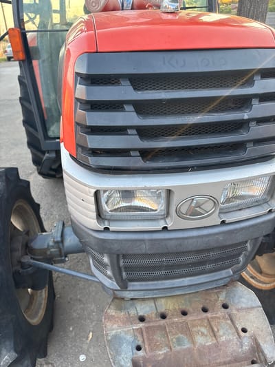 A close-up, low-angle shot captures the front of a red and gray Kubota tractor parked outdoors on a paved surface. The tractor is the primary subject. The front grille is prominent, with horizontal black slats over a finer mesh. The word "Kubota" is visible in gray lettering on the upper black section of the grille. Below the grille are two rectangular headlights, one on each side, with clear lenses and visible internal components. A silver oval emblem with a stylized "K" is centered between the headlights. The tractor's front tire, a large black rubber wheel with a thick tread, is visible on the left side of the frame. A metallic gray steering linkage extends from the tractor's chassis towards the wheel. The bottom portion of the tractor reveals a rusted metal platform with several circular holes. The background is blurred, showing the right side of the tractor's cabin with an orange indicator light and tinted windows. Hints of trees and what appears to be a residential building with a car are visible in the far background, suggesting an outdoor, possibly semi-rural or suburban setting. The lighting indicates it is daytime, with some sunlight glinting off the tractor's surfaces. No people are visible in the image. The scene is static and shows the tractor parked.