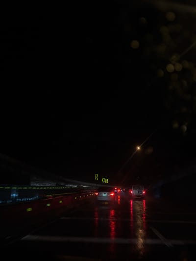 The image depicts a nighttime scene from inside a vehicle on a wet multi-lane highway in San Lorenzo Acopilco, Mexico. The environment is predominantly dark, with a dark overhead structure suggesting an underpass or elevated section of road. The asphalt is visibly wet, reflecting the red taillights of multiple vehicles ahead, creating distinct light streaks on the road surface.
Essential details include:
* **Vehicles:** A light-colored sedan is visible in the left-center lane, and a white van is present in the right lane, both displaying illuminated red taillights. Additional vehicle taillights are visible further down the road.
* **Road Infrastructure:** The left side of the highway is bordered by a series of red modular barriers, each equipped with horizontal reflective strips. White dashed lane markings are discernible on the wet road.
* **Signage:** Two yellow chevron directional signs are mounted overhead in the mid-ground.
* **Lighting:** Beyond vehicle taillights, a single distinct point of light, likely a distant streetlight, is visible in the upper mid-right. Blurred, circular light sources are present in the top right quadrant, consistent with reflections or water droplets on a windshield.
* **Actions:** Vehicles are proceeding along the wet highway. No individuals are visible outside the vehicles.