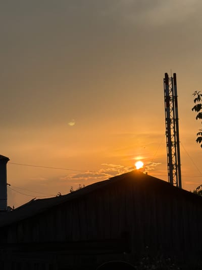A silhouetted view of buildings and a tall industrial structure against a dusky sky in Vikulovo, Russia, as the sun sets. The sun, a bright orb, is partially obscured by the peak of a dark, wooden roof in the foreground. To the right, a tall, metallic tower with red and white markings rises vertically. The sky is a warm gradient of orange and yellow, with scattered clouds. A dark branch with leaves is visible on the far right. The image captures a tranquil yet slightly industrial atmosphere, characteristic of a small town at dusk.