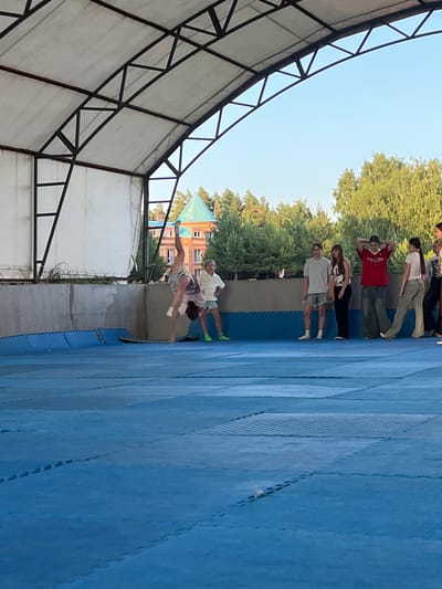 In an outdoor setting in Krasnoturansk, Russia, a group of young people are engaged in what appears to be a gymnastics or acrobatic training session. The scene is set on a bright, clear day, with a blue sky visible above. The ground is covered with large, interlocking blue mats, typical of a training facility.
A structure with a white canopy and a metal frame is visible to the left, suggesting an outdoor training area or a covered section of a larger facility. In the background, lush green trees and a building with distinctive green-tiled roofs and brickwork provide context to the location.
Several individuals are captured in various poses. One person is performing an acrobatic move, with their body in a dynamic, arched position, legs extended. Another individual, wearing a white shirt and shorts, appears to be assisting or spotting them. Further back, a line of people are standing, some in casual athletic wear, watching the activity. Their stances suggest they are either waiting for their turn, observing, or possibly participating in warm-up exercises. The overall mood seems to be one of active training and camaraderie among the participants. The time of day is likely afternoon, given the bright lighting and the clear sky.
