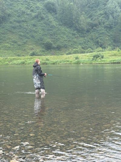 A person stands in the shallow water of a river, holding a fishing rod. They are wearing waders and a dark, hooded jacket with white markings. The riverbed is visible, covered in stones. In the background, a lush, green hillside rises steeply, covered in trees and vegetation. The sky appears overcast. The location is the city of Ust-Kabirza, Russia.