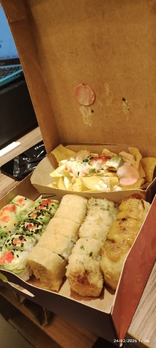 An open, brown cardboard takeout box, presumably containing food from a commercial establishment in Tumbaco, Ecuador, is presented from a slightly elevated, angled perspective. The box is divided into sections. The largest section, occupying the lower half, contains multiple rows of sushi rolls. From left to right, these include sushi rolls topped with green avocado slices, red roe, and black sesame seeds; several sections of light-colored, possibly tempura-fried, sushi rolls; and an additional section of similar light-colored rolls topped with a caramelized or glazed substance, potentially plantain or banana. The upper right section of the box contains a smaller cardboard tray filled with yellow chips, consistent with plantain or corn chips, topped with a white sauce, diced green and red vegetables (likely avocado and tomato), green chives, and a pink, circular piece of an unknown food item. The interior of the box lid displays smudges and a light pink circular piece, possibly processed meat or candy. The background shows a partial view of a wooden surface, a dark object (possibly a phone or wallet), a white straw or stick, and a portion of a laptop screen displaying a blue interface. A timestamp "24/02/2026 17:36" is visible in the bottom right corner of the image. No individuals are present, and no active interactions are depicted.