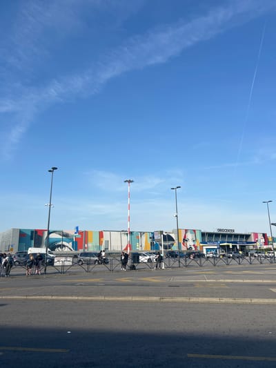 The image depicts the OrioCenter shopping mall in Orio al Serio, Italy, on a clear day with a bright blue sky and wispy clouds. The facade of the mall features a large, colorful mural with abstract designs and stylized human faces. In front of the mall, a street is visible with a black metal fence separating it from a sidewalk where several people are standing, some with luggage. Cars are parked in front of the mall and driving on the street. Tall lampposts with multiple lights are positioned throughout the scene. A red and white striped pole stands near the right side of the image. The ground is paved, with yellow lines marking lanes on the street.