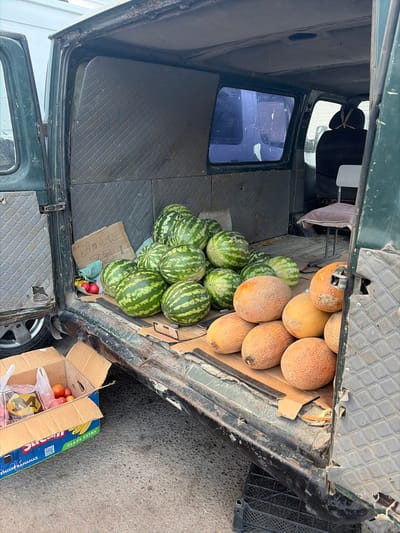 The image depicts the interior and exterior of an open vehicle, possibly a truck or van, filled with various fruits. In the back of the vehicle, a collection of cantaloupe melons with textured orange skin are piled up. Behind the melons, several watermelons with dark green stripes are stacked. Some of the watermelons are partially obscured by other fruits or the vehicle's interior. A small, worn cardboard sign with what appears to be handwritten text in a non-Latin script rests among the watermelons. Two red fruits, possibly apples or tomatoes, are also visible near the bottom of the watermelon pile.
To the right of the vehicle, a cardboard box lies open on the ground. The box is labeled with text, including "PREMIUM BANANAS" and possibly other branding information in a blue and yellow color scheme, suggesting it may have previously contained bananas or is intended for fruit transport. Inside the box, several red fruits, likely tomatoes or apples, are visible along with a plastic bag containing items that are difficult to discern.
The vehicle itself appears weathered and utilitarian, with a dark green exterior and a worn interior featuring textured gray paneling. The open door of the vehicle reveals a glimpse of the driver's side, including a seat and a window with a blue tint. The setting appears to be outdoors, on a paved or asphalt surface, suggesting a market or roadside stall. The lighting indicates it is daytime. The overall scene suggests a commercial activity, likely the sale or transport of fresh produce. There are no people visible in the image.