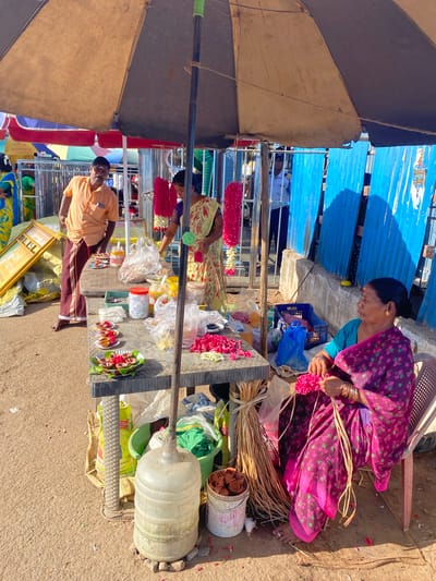 The image depicts an outdoor scene in Chinnambedu, India, beneath a large, weathered umbrella. A vendor, a woman in a vibrant purple sari adorned with pink floral patterns, sits in a chair on the right, meticulously crafting flower garlands. She holds a bundle of pink flowers and dried grass stems, her hands actively engaged in the creation process. To her left, a table displays various items, including small clay diyas (lamps), flower petals, jars of what appear to be food or offerings, and plastic bags. Another vendor, a man wearing an orange shirt and a traditional lungi, stands behind the table on the left, looking towards the camera. Behind the vendors, a blue corrugated metal fence forms a backdrop. Further in the background, glimpses of other stalls, colorful fabrics, and people suggest a bustling market or festival environment. The ground is composed of dirt and scattered flower petals. A large, semi-transparent water dispenser is visible on the lower left, next to a green bucket and sacks. The lighting suggests it is daytime, with strong sunlight casting shadows.